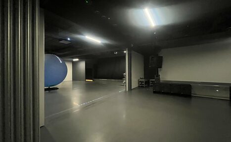 Арендовать Futurione(Футурио́не). Просторное помещение с гладким серым полом, темными стенами и потолком, освещенное потолочными светильниками. Слева виден большой синий объект. Справа расположено звуковое оборудование и черные диваны.