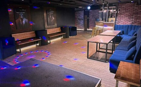 Арендовать BULL LOFT(Лофт Бык) . Лофт-пространство с кирпичными стенами, деревянным полом и баром. Угловой диван и скамьи вдоль стен, длинные деревянные столы. Потолочное освещение, неоновая подсветка и световые пятна на полу. Два постера на стене.
