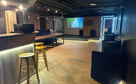 Арендовать BULL LOFT(Лофт Бык) . Современное помещение с деревянными полами и кирпичными стенами. Барная стойка с высокими стульями, черные кресла, проектор с изображением на экране, направленное освещение на потолке, деревянные панели, светлое и просторное пространство.