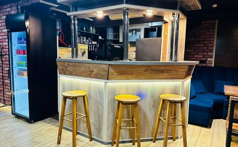 Арендовать BULL LOFT(Лофт Бык) . Барная стойка с деревянной отделкой, три деревянных стула. За стойкой полки с напитками и оборудованием. Справа тёмно-синий диван и деревянный стол. Светлое деревянное покрытие пола, кирпичная стена, холодильник с напитками слева.