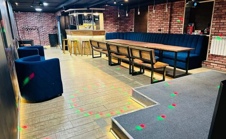 Арендовать BULL LOFT(Лофт Бык) . Арендуемое пространство с барной стойкой; кирпичные стены; деревянные полы и барные стулья; длинный стол с лавкой и синими креслами; черный потолок со светильниками; разноцветные световые точки на полу.