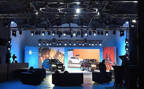 Арендовать Studio Hall(Студио Холл). Сцена с двумя автомобилями, освещённая яркими прожекторами и окружённая синим светом. В помещении видны конструкции с тросами и софитами. На заднем плане крупные экраны с изображениями автомобилей. Впереди мягкие чёрные кресла.
