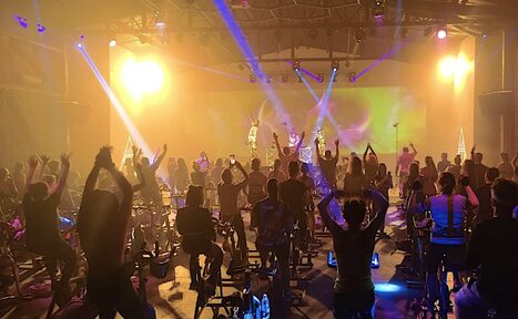 Арендовать Studio Hall(Студио Холл). Атмосферное помещение для групповых тренировок, расставлены велотренажеры, много людей занимается. Яркое освещение, световые эффекты с прожекторами, виден экран. Потолок с видимыми балками, полы тёмного цвета, индустриальный стиль.