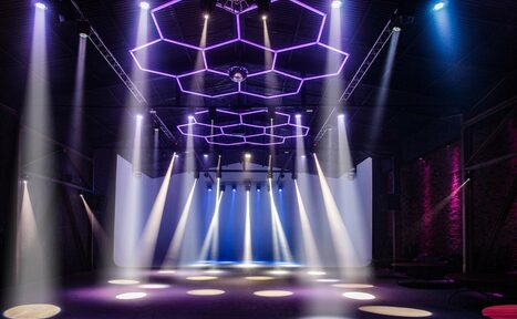 Арендовать Studio Hall(Студио Холл). Просторное помещение с черным потолком и кирпичными стенами. Видна сцена с белыми занавесями. Световое оборудование создает разноцветные световые пятна и линии, включая сиреневые и синие оттенки. Потолок декорирован шестигранниками.
