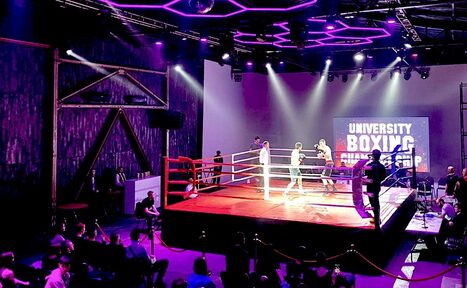 Арендовать Studio Hall(Студио Холл). Изображение показывает спортивное пространство с боксерским рингом в центре. Вокруг ринга собрались зрители. Зал освещен фиолетовыми и белыми прожекторами. На стене экрана видна надпись "University Boxing Championship".