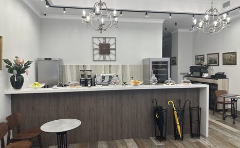 Арендовать Арт Холл. Кафе с барной стойкой из дерева, белой столешницей. На стойке кофемашина, вазы с фруктами и десертами. Сзади холодильник и шкаф. Светильники на потолке, картины на стенах. Деревянные пол и стулья, столы с мраморными столешницами. Зонты за стойкой.