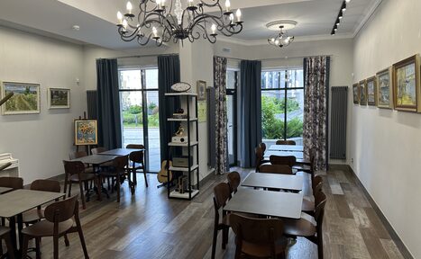 Арендовать Арт Холл. Небольшое помещение с кафейными столами и стульями, стены украшены картинами. На полу деревянный ламинат, освещение от современных и классических люстр. Большие окна с плотными шторами. Справа книжная полка с декорациями.