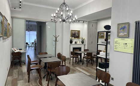 Арендовать Арт Холл. Комната с деревянными столами и стульями, белые стены с картинами, подвесная люстра. Стеклянная дверь ведет в соседнее помещение. Пол из светло-коричневого дерева. В углу стоит вешалка, у стены камин. На стене план помещения.