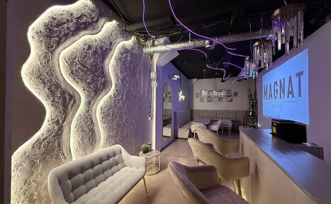 Арендовать Лофт SUNRISE(Восход Солнца). Просторное помещение на 15 гостей. Современное помещение с приглушенным освещением, мягкими креслами и диванами, декоративной подсветкой на стенах. На стене знак "MAGNAT". Видно барную стойку, подвесные светильники, трубы на потолке, картины на стене.