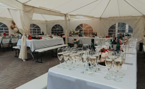 Арендовать Усадьба Афонинская. Белый шатёр с боковыми стенками из ткани и пластиковыми окнами. Внутри расставлены накрытые белыми скатертями столы с напитками и бокалами, деревянные лавки. Освещение естественное, пол выложен плиткой.