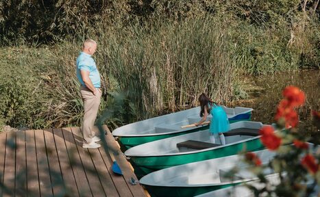 Арендовать Усадьба Афонинская. Деревянный причал у воды с тремя зелеными лодками, одна из которых занята ребенком. Взрослый стоит на причале. Вокруг густая растительность и цветы.