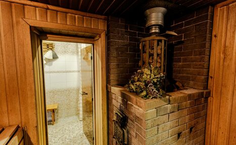Арендовать Усадьба Афонинская. Сауна с деревянной отделкой, справа кирпичная печь с камнями и металлическим дымоходом. Слева стеклянная дверь, за ней светлая комната с белыми плитками и стеной из мелких камней, деревянная скамья. Освещение тёплое.