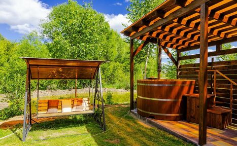 Арендовать Усадьба Афонинская. Открытое пространство с деревянной беседкой и деревянной купелью, справа деревянная терраса с перголой. Слева подвесные качели с подушками. Поверхность из травы и дерева, фон из деревьев. Яркое дневное освещение.