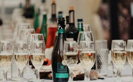 Арендовать Усадьба Афонинская. Стол с бокалами для вина и бутылками на фоне, в центре бутылка игристого вина с зелёной этикеткой, бокалы частично наполнены белым вином, вокруг другие стеклянные и зелёные бутылки, белая скатерть под ними.