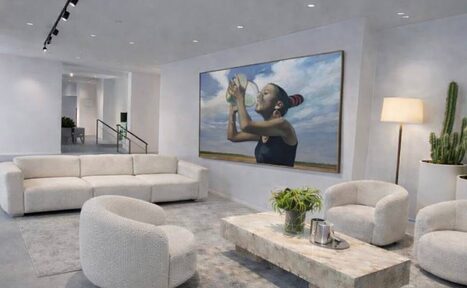 Арендовать Level 5(Лэвел 5). Современное пространство с мягким диваном и креслами белого цвета, центральный стол из светлого камня. На стене картина с изображением человека. Напольная лампа и кактус в углу. Светлый ковёр, потолочные светильники.