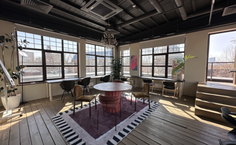 Арендовать Пространство с террасой до 50 человек. Просторная комната с деревянным полом, круглым столом и стульями. Большие окна с естественным светом, люстра, растения, ковёр с узором, картины на стенах. Черный потолок с трубами и кондиционерами, мягкие кресла у стен.