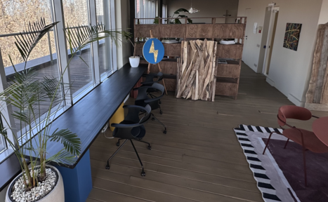 Арендовать Пространство с террасой до 50 человек. Современное офисное пространство с длинным столом, черными офисными креслами, деревянными перегородками. Большие окна, много естественного света. Плоские растения в горшках, деревянный пол, цветная мебель, декоративная зелень, абстрактная картина.