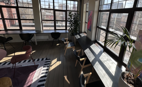 Арендовать Пространство с террасой до 50 человек. Лофт-пространство с деревянным полом и большими окнами. Вдоль окон столы с черными стульями. Растения в горшках. На полу ковер с геометрическим узором. Красно-кирпичная стена видна через окна.