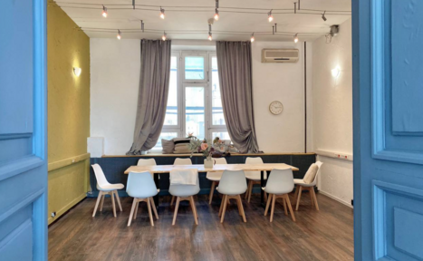 Арендовать H4U. Светлый зал П-602. Просторная комната с деревянным полом, большим светлым столом и белыми стульями. На стене часы, рядом окно с серыми шторами. Подвесные лампы на потолке, кондиционер на стене. Светлая и аккуратная атмосфера.