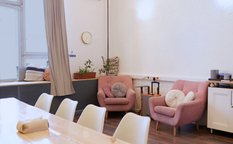 Арендовать H4U. Светлый зал П-602. Просторная комната с деревянным столом и белыми стульями. В углу находятся два розовых кресла с декоративными подушками и небольшой столик. На стене часы, на подоконнике подушки и растения. Светлое освещение, белые стены, деревянный пол.