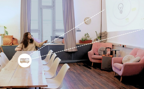 Арендовать H4U. Светлый зал П-602. Просторное помещение с деревянным столом и белыми стульями; на стене проекция изображения. В углу розовые кресла и подушки, между которыми небольшой столик и динамик. Пол тёмный деревянный. Есть окно с занавесками, комнатные растения и настенные часы.