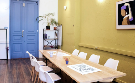 Арендовать H4U. Светлый зал П-602. Просторная комната с деревянным столом для собраний, окружённым белыми стульями. Слева дверь синего цвета, на стене зелёного оттенка светлое бра и картина. Пол тёмного дерева, на столе раскраска и цветные карандаши, растения на полке.