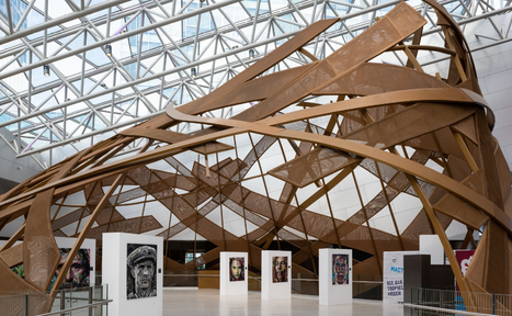 Арендовать Кунцево Плаза. Выставочное пространство с витиеватой структурой из коричневых деревянных элементов под стеклянной крышей. На белых стендах расположены портретные картины. Белый пол, металлические перила, знаки с текстами на заднем плане.
