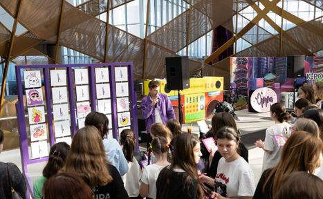 Арендовать Кунцево Плаза. Мероприятие в помещении с высокими окнами и геометрическими конструкциями на потолке. Множество людей смотрят на стенды с изображениями персонажей. Есть микрофон и колонки. Яркие цвета: фиолетовый, жёлтый, розовый.