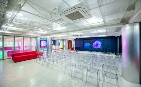 Арендовать АКИ.лаб пространство для мероприятий. Просторное современное помещение в стиле конференц-зала с серыми стенами и потолком из перфорированных панелей. Прозрачные стулья расположены рядами перед большим экраном на стене. Слева красный диван. Пол светлый, интерьер освещен ярким искусственным светом.