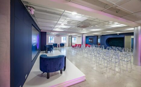 Арендовать АКИ.лаб пространство для мероприятий. Современное конференц-пространство с панорамной планировкой и сценой, прозрачные стулья для зрителей, мягкие кресла на сцене. Большой экран, ровное освещение, бетонный пол, белый и синий цвета интерьера.