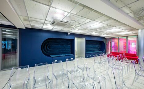 Арендовать АКИ.лаб пространство для мероприятий. Современное помещение со стеклянными стенами и прозрачными стульями. Синяя облицовка стен с нишами, белый потолок с подсветкой, кондиционер. Напольное покрытие светлое, вдоль стены красные диваны. Пространство предназначено для мероприятий.