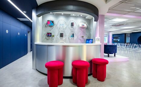 Арендовать АКИ.лаб пространство для мероприятий. Современное помещение с серебристой стойкой и тремя красными круглыми табуретами. На стене за стойкой круглые ниши с предметами. Пол бетонный, стены темно-синие. Освещение яркое, со встроенными светильниками.