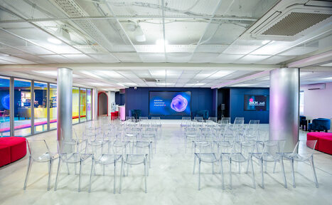 Арендовать АКИ.лаб пространство для мероприятий. Аудиторное пространство с прозрачными пластиковыми стульями, расположенными рядами. Светлые стены и потолок с встраиваемым освещением. Две металлические колонны. На стене мониторы с изображениями и текстом. Пол светлый, глянцевый.