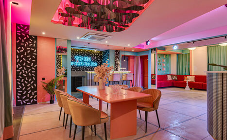 Арендовать Лофт Шекспир. Стильный дизайнерский зал. Современное пространство с открытой планировкой. В центре розовый стол и стулья, барная стойка с высокими стульями, разноцветные мягкие диваны, неоновая вывеска. Светодиодное освещение, разноцветные стены и декор, пол из светлых плит.