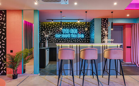 Арендовать Лофт Шекспир. Стильный дизайнерский зал. Барная зона с высокой стойкой и четырьмя барными стульями. На фоне неоновая надпись "TO BE or not to be". Стены декорированы черно-белыми узорами. Освещение подвесное, теплое. Видно растение в горшке и занавеску.