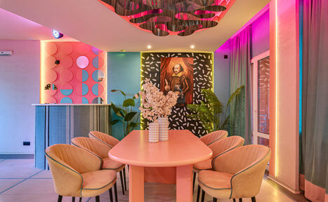 Арендовать Лофт Шекспир. Стильный дизайнерский зал. Пространство с длинным розовым столом и бежевыми стульями, декор с цветами на столе. Яркое освещение с розово-фиолетовыми неоновыми акцентами. На стенах постер с портретом, искусственные растения, шторы и барная стойка с круглым узором.