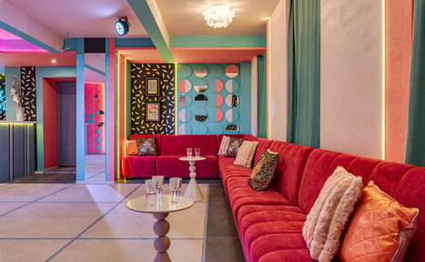 Арендовать Лофт Шекспир. Стильный дизайнерский зал. Просторная комната с длинным красным диваном вдоль стены, разноцветными декоративными панелями на стене, круглыми столиками с прозрачными стаканами, мягкими подушками. Яркое освещение, плиточный пол, разноцветные стены в геометрическом стиле.