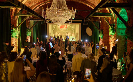 Арендовать Arsenal Loft(Арсенал Лофт). Арочный зал с кирпичными стенами, большим кристальным люстрой и деревянными балками. Пространство заполнено людьми, на сцене группа. На заднем плане светящийся экран с надписью "AURA". Столы с белыми скатертями и прозрачными стульями.