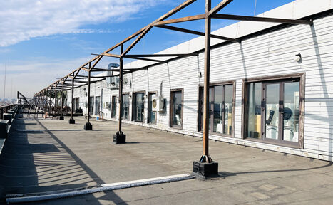 Арендовать Зал под кулинарные мастер классы и мероприятия. Просторная открытая терраса с бетонным полом вдоль здания с белой деревянной облицовкой. Металлические столбы поддерживают конструкции над террасой. Большие окна с рамами, несколько кондиционеров на стене. Яркое дневное освещение.