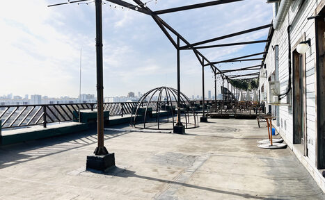 Арендовать Зал под кулинарные мастер классы и мероприятия. Открытая терраса на крыше с бетонным полом, металлическими перголами и ограждением. Справа вдоль стены установлены светильники. По центру пустая металлическая конструкция. На горизонте виден городской пейзаж.