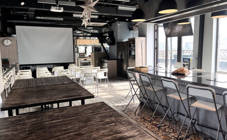 Арендовать Зал под кулинарные мастер классы и мероприятия. Пространство с проектором и экраном, множеством белых стульев и деревянными столами. Барная стойка с высокими стульями справа, большие окна, тёмные подвесные светильники, деревянный и плиточный пол. Потолок с открытыми коммуникациями.