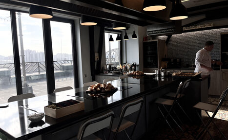 Арендовать Зал под кулинарные мастер классы и мероприятия. Кухонное пространство с длинным столом-островом, черной столешницей, белыми стульями и приборами. Широкие окна с видом на город. Подвесные светильники, кирпичная стена, плитка на полу. На столе булочки и кофеварка.