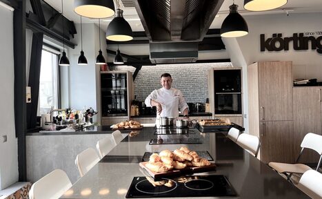 Арендовать Зал под кулинарные мастер классы и мероприятия. Кухонное пространство с несколькими индукционными плитами на серых столах, стульями с белыми сиденьями. Над столами подвешены черные и белые лампы. На заднем плане, повар готовит еду в кастрюле. Справа видны шкафы и полки.