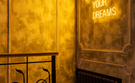 Арендовать Каминный Зал Tangiers lounge Pokrovka(Танжерс Лаундж Покровка). Золотистая стена с декоративными элементами, черный радиатор. Металлические перила слева внизу. Яркая неоновая надпись на английском языке. Теплое освещение.