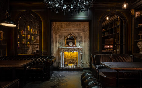 Арендовать Tangiers lounge Pokrovka (Танжерс Лаундж Покровка). Каминный Зал . Тёмное лаунж-пространство с кожаными диванами и деревянными столами. Центрально располагается декоративный камин с зеркалом и свечами. Арочные окна, полки с книгами, бюст. Освещение жёлтое, мягкое, с крупной чёрной люстрой.