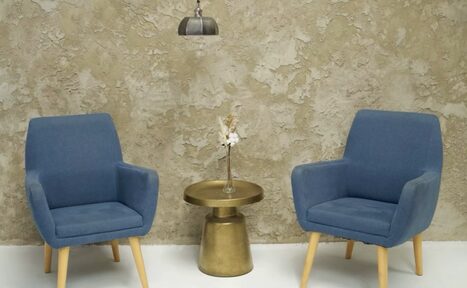 Арендовать WhiteFox Studio(Студия Белая Лиса). Две синие кресла с деревянными ножками стоят по бокам круглого золотистого столика с вазой на фоне декоративной стены с фактурной отделкой. Над столиком подвешен металлический светильник. Пол светлый.