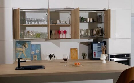 Арендовать WhiteFox Studio(Студия Белая Лиса). Кухонное пространство с деревянной столешницей, открытые шкафы со стеклянной и керамической посудой, картина, ваза на столе, черный кран, бокал с напитком, тарелка с едой, встроенная духовка, светодиодное освещение, нейтральные оттенки.