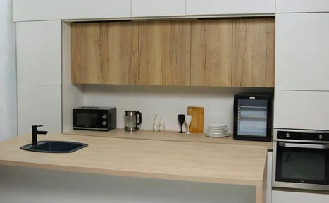 Арендовать WhiteFox Studio(Студия Белая Лиса). Современная кухня с деревянной столешницей и шкафами. Есть встроенная духовка, микроволновка, чайник, раковина с краном, холодильник, стеклянные бокалы, стопка тарелок, разделочная доска. Декор светлый, с деревянными и черными акцентами.