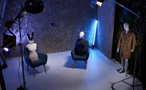 Арендовать WhiteFox Studio(Студия Белая Лиса). просторная студия с серыми текстурированными стенами, двумя синими креслами, на которых стоят манекены, справа манекен в пальто, освещение студийное с цветными акцентами, пол светлый, в углу неоновая лампа, растительность у стены справа.