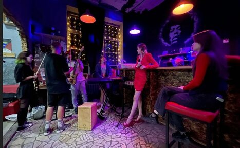 Арендовать Лофт-Бар. Музыкальное пространство с барной стойкой, на стене портрет музыканта. Группа людей с инструментами, включая клавиши и саксофон. Красные и фиолетовые подвесные светильники, фонари за окном. Плиточный пол, разноцветная мебель.